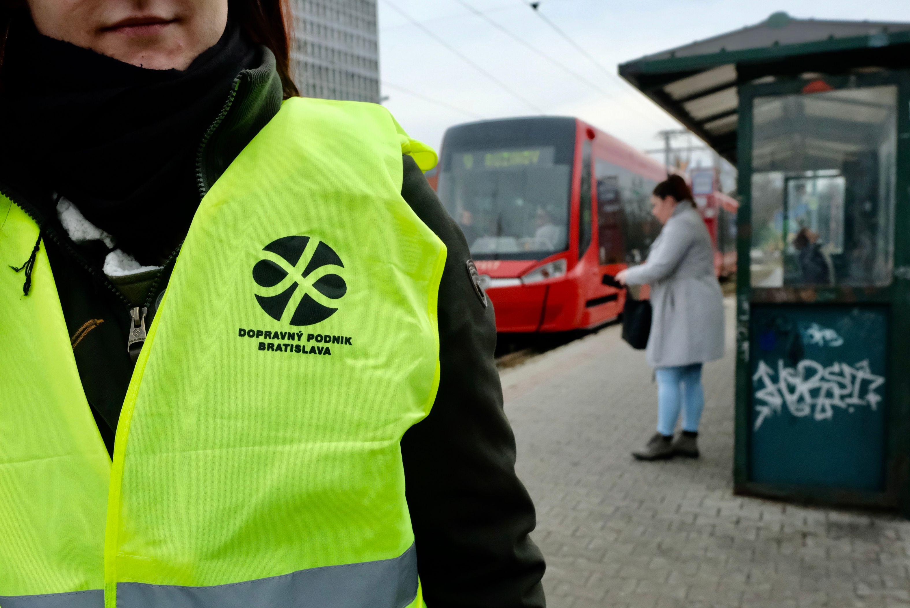 človek s reflexnou vestou dopravného podniku stojí na zastávke, v pozadí je nová bratislavská električka